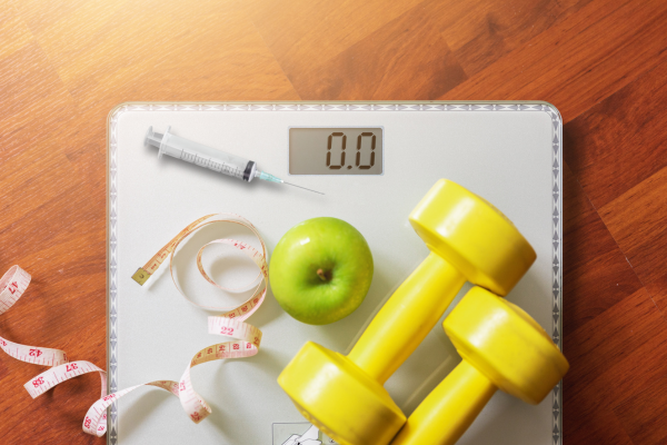 An image of a scale with a syringe for injectable vitamins, an apple, dumbbells, and a measuring tape
